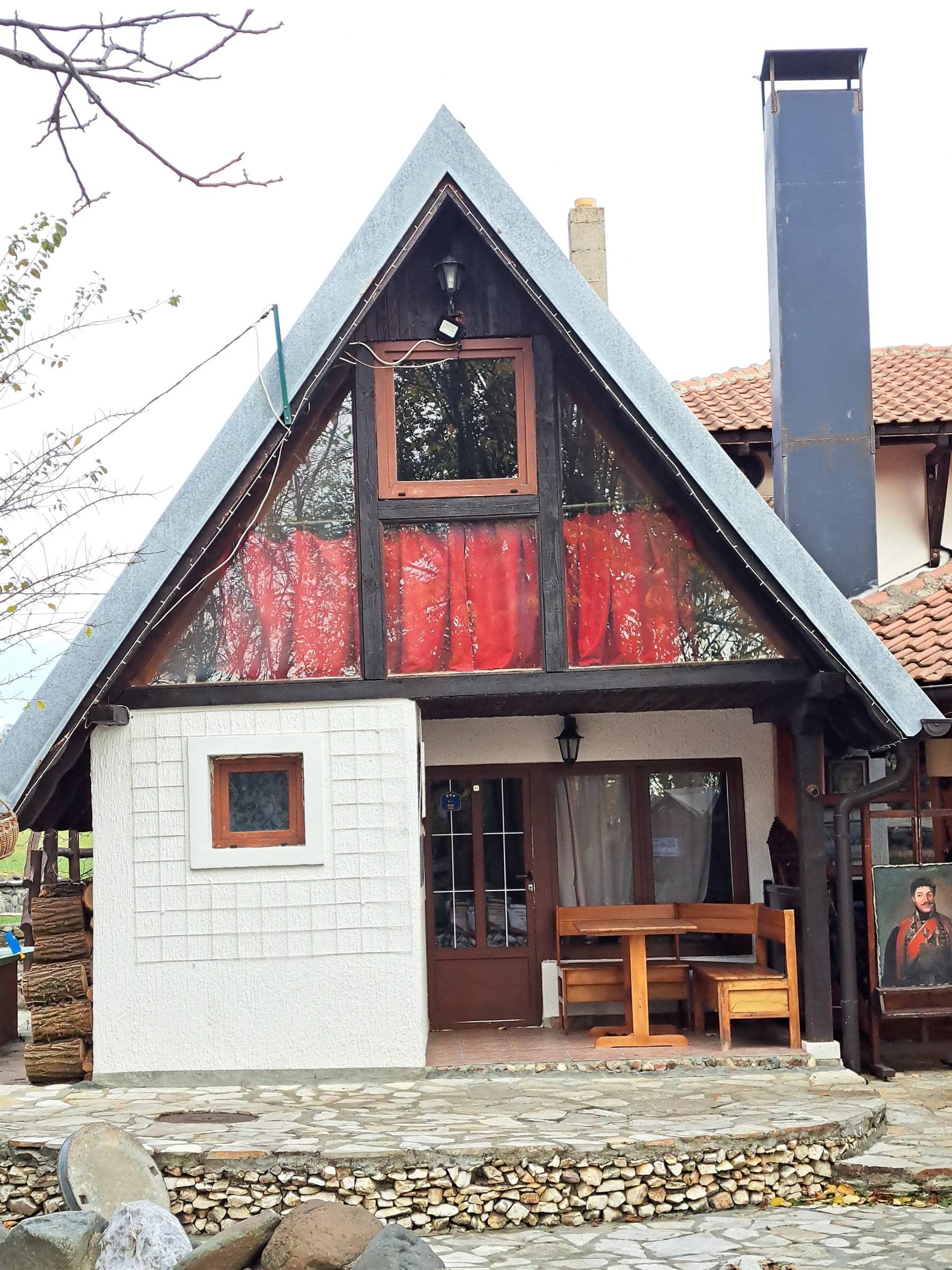Bungalov 1: kućica sa strmim krovom, crvenim zavesama na spratu i stolom ispred ulaza