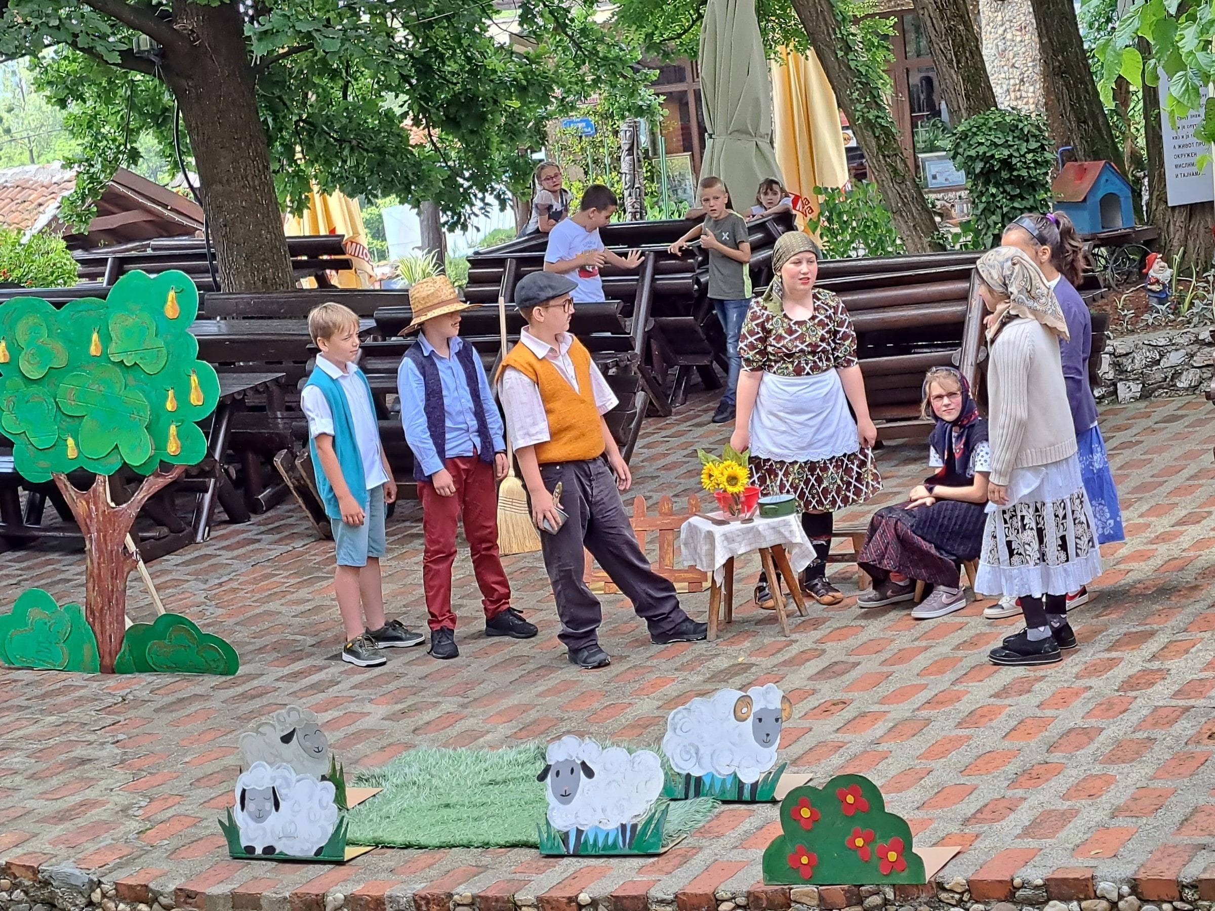 Deca u narodnim nošnjama izvode predstavu u dvorištu, ispred drvenih ovaca